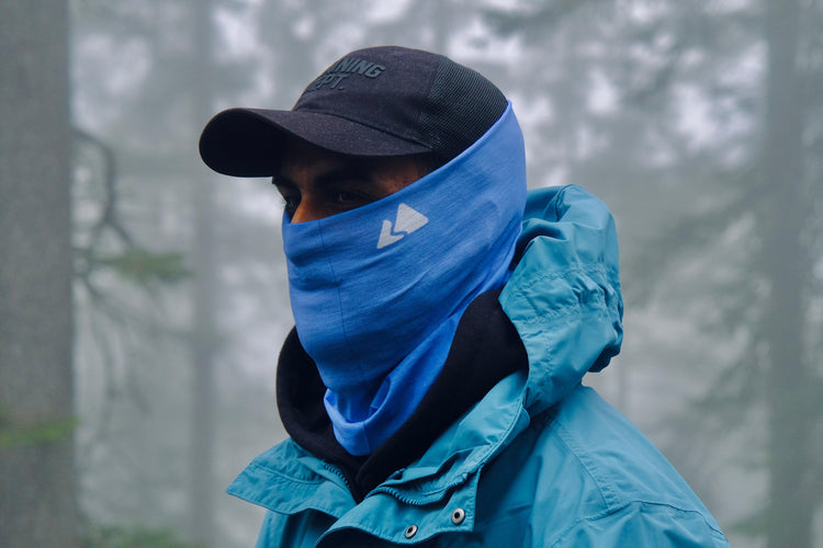 Day sky blue hiking buff snood with a white mountain logo in the left hand side between the ear and the mouth. Protecting against the fog and wind. Model wearing black hat and blue windbreaker jacket 