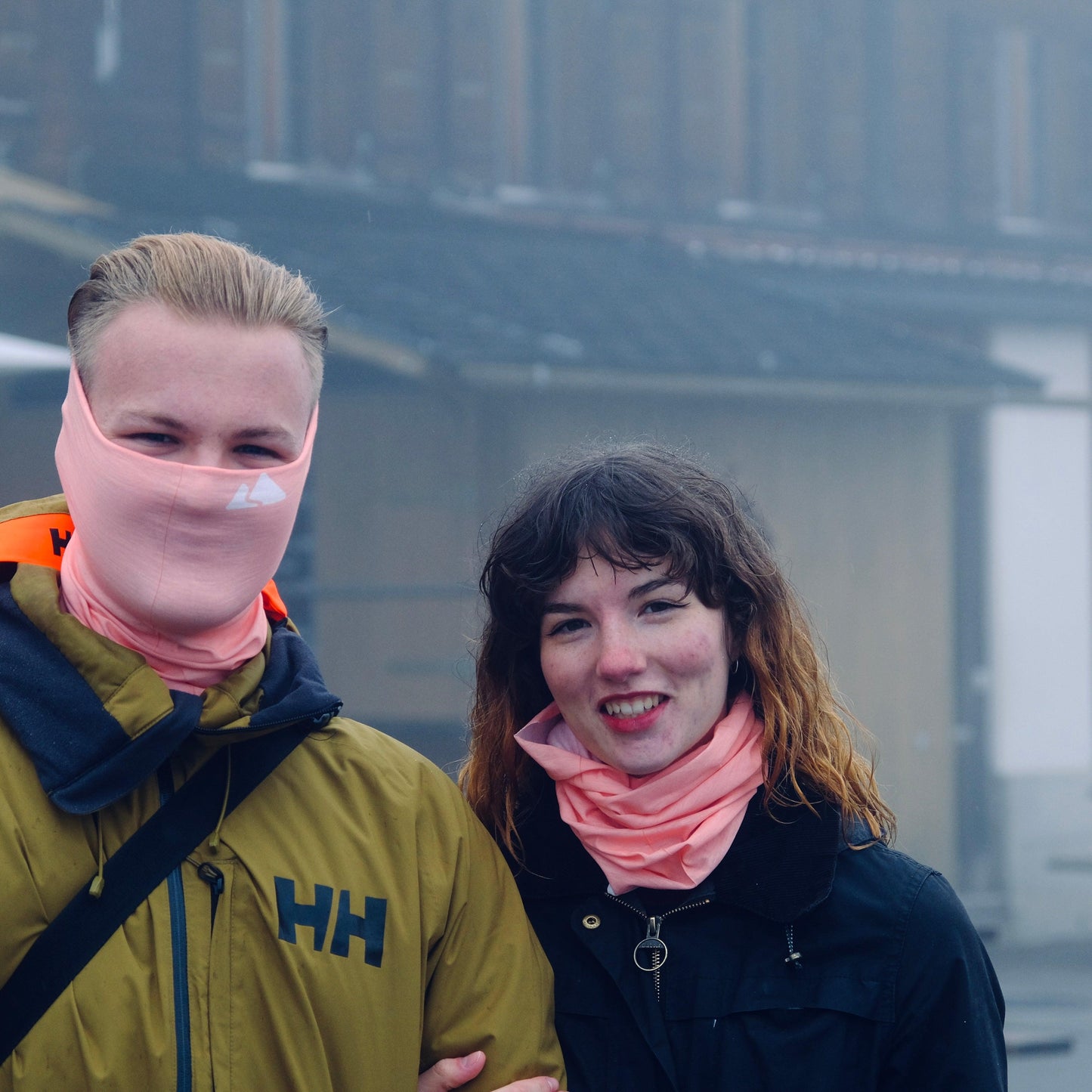 Neck buff gaiter displayed in a male and female model. With a white mountain logo. Blush Peach colour.  